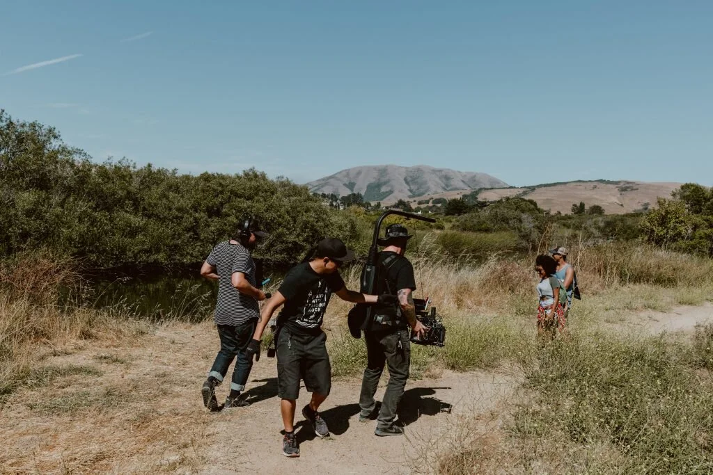 Film crew filming a scene outdoors in a rural area near shrubbery and a body of water, with a mountain in the background, under a clear blue sky.