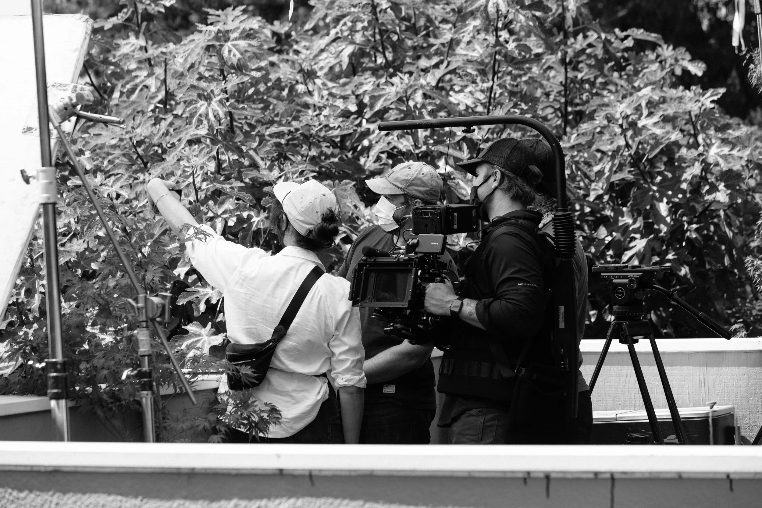 A black and white photo of a film crew filming a scene in a lush, leafy outdoor setting. Three crew members are wearing face masks and caps, focusing on their work with cameras and equipment.