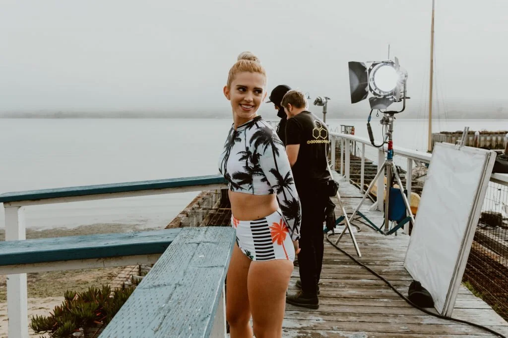 A young woman in a tropical-themed swimsuit and cover-up standing on a dock near the water, with a film crew setting up lighting equipment behind her.