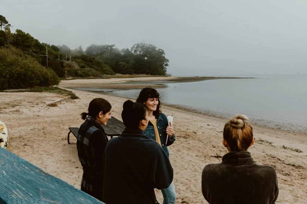 Four people standing and talking on a sandy beach, with trees and calm water in the background on a foggy day.