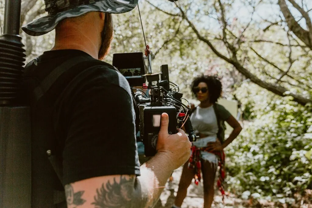 A person operating a camera outdoors, filming a woman posing with hands on hips in a wooded area.