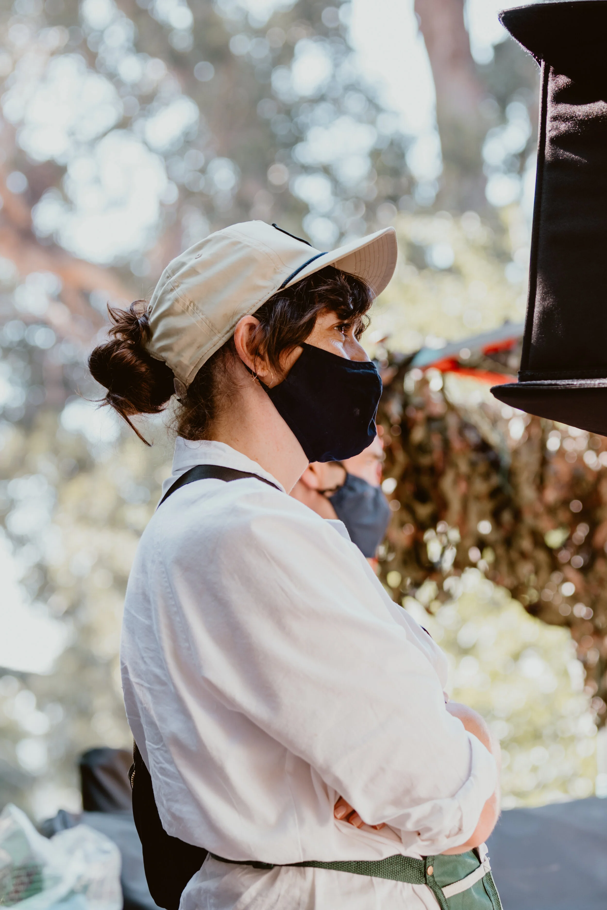 A woman wearing a beige cap and black face mask outdoors, with her arms crossed. She has dark curly hair tied in a bun and is dressed in a white jacket. There are blurred trees and people in the background.