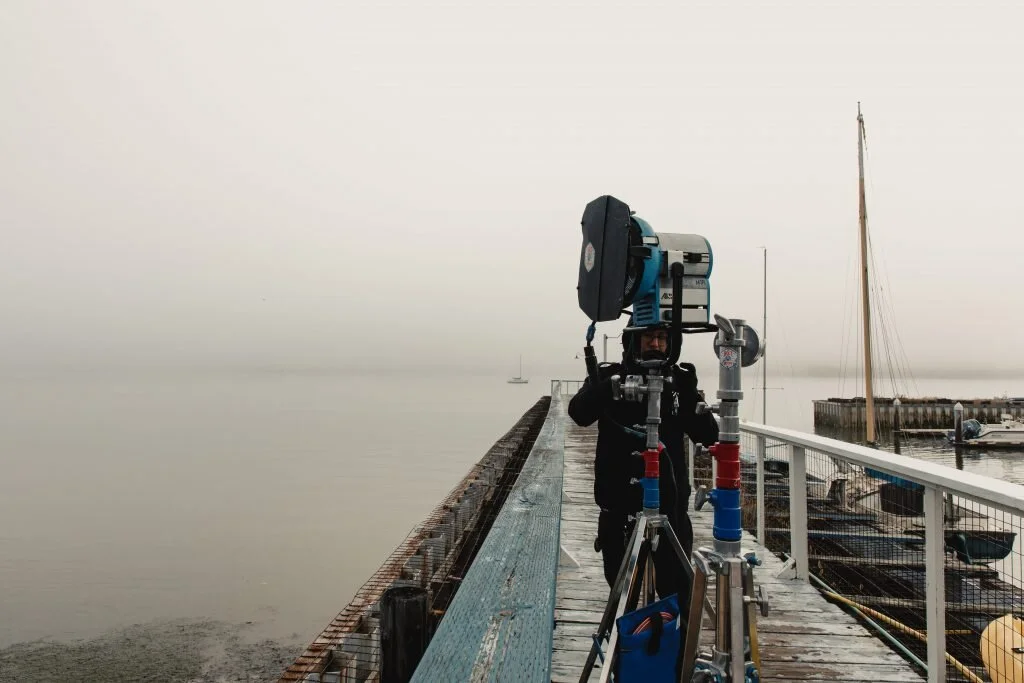 Person operating telescope equipment on a harbor pier with sailboats and a foggy coastline in the background.