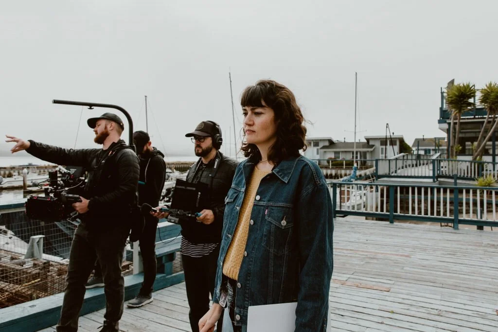 A woman stands outdoors on a wooden deck near a marina, with a group of filmmakers filming a scene nearby.