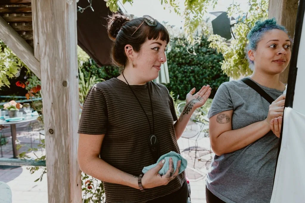 Two women outdoors, one with dark hair in a bun, wearing a striped black and brown shirt, holding a face mask, gesturing with her hand, and talking to the other woman with blue hair, wearing a gray shirt, standing near a white backdrop.