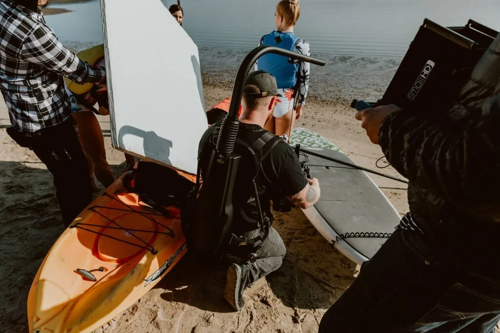 Group of people preparing sailboards and a paddleboard by the water at the beach, with one person kneeling and adjusting equipment while others stand nearby.
