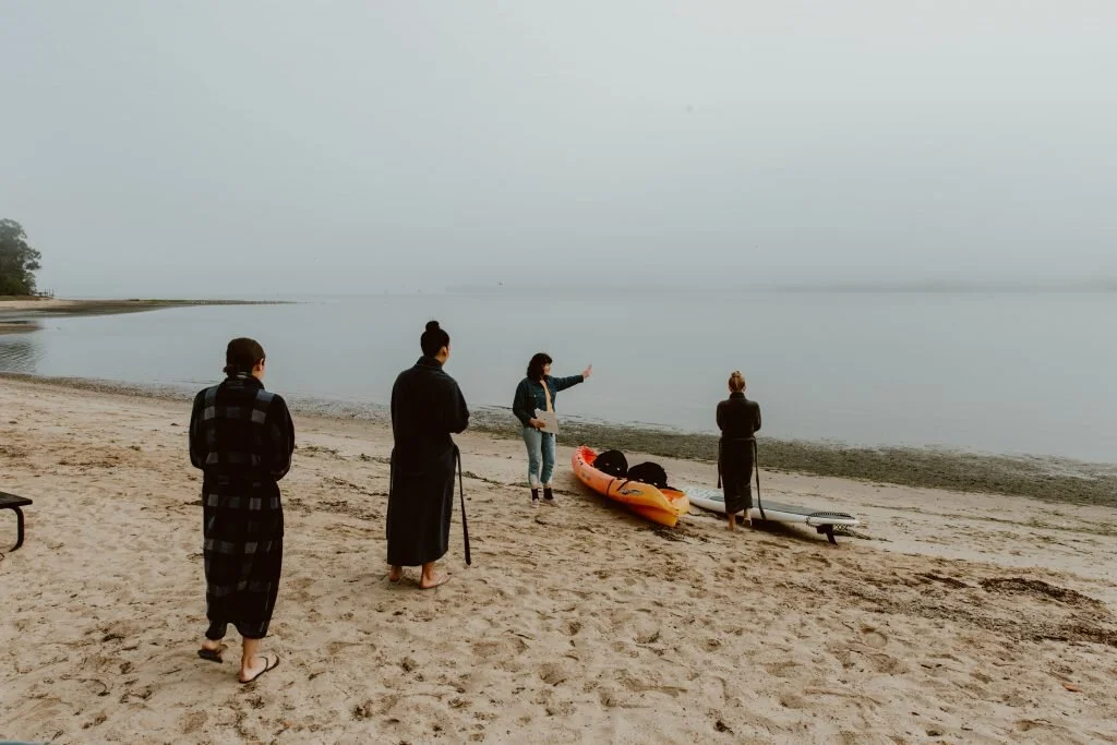 Four women stand on a sandy beach near a boat and a paddleboard, facing calm water with a cloudy sky, engaging in conversation or instruction.