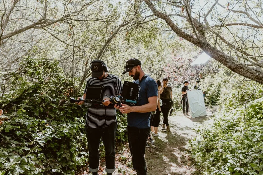 Two filmmakers on a forest trail operating a camera and monitor with a small crew working in the background, set up for filming outdoors.
