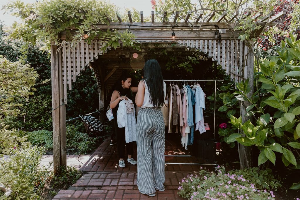 Two women shop for clothes at an outdoor market stall surrounded by greenery.