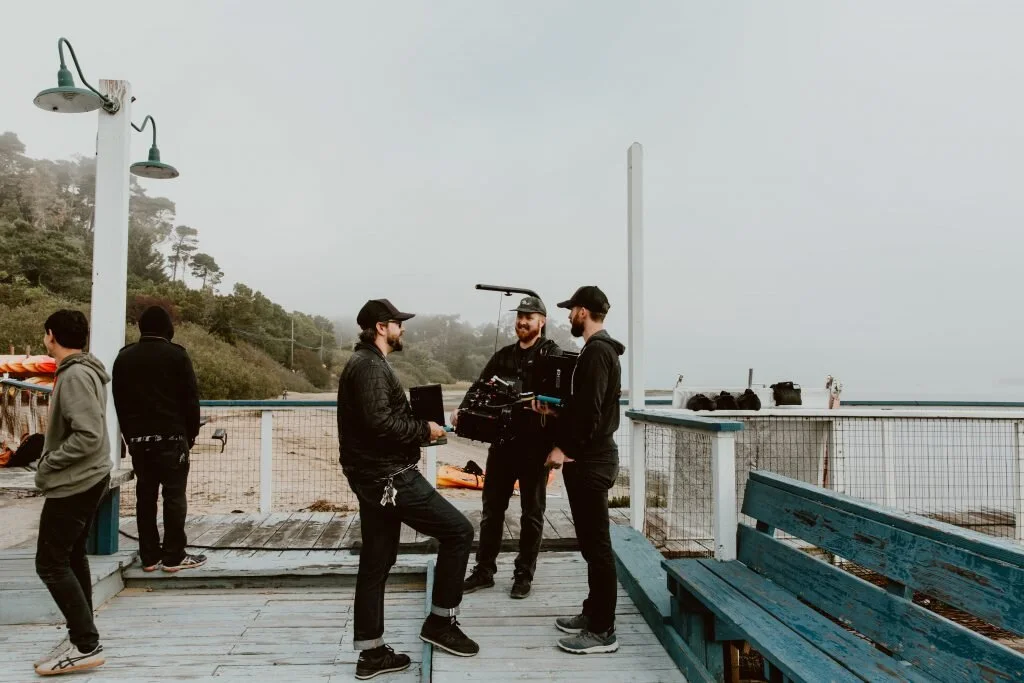 Group of five men standing and talking on a wooden pier near the beach, with ocean and foggy hills in the background.