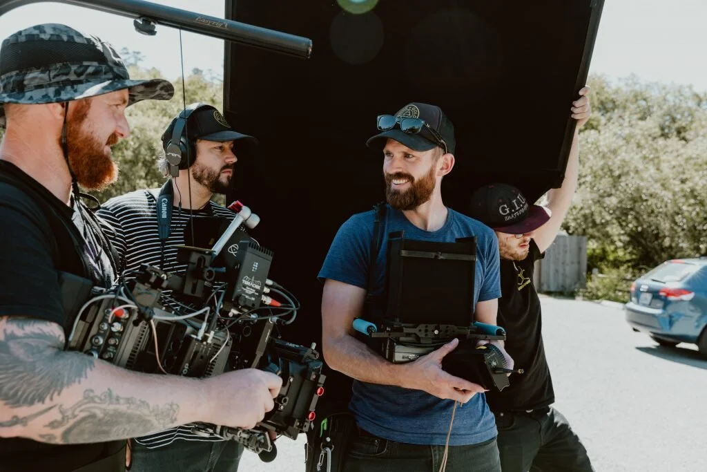 A group of four men working together outdoors with film equipment, under a large black diffuser or reflector, on a sunny day.