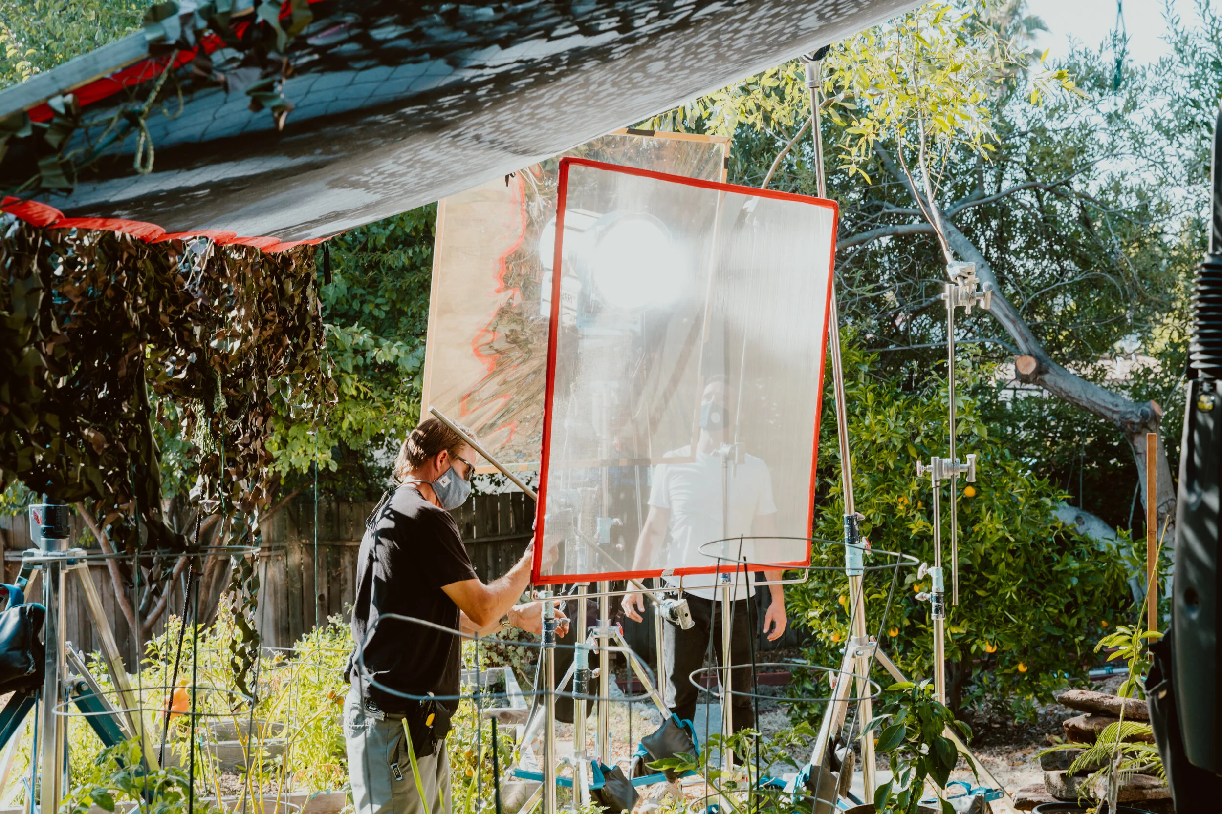 Filmmakers set up lighting and equipment outdoors in a garden with trees and orange trees, preparing for a shoot.