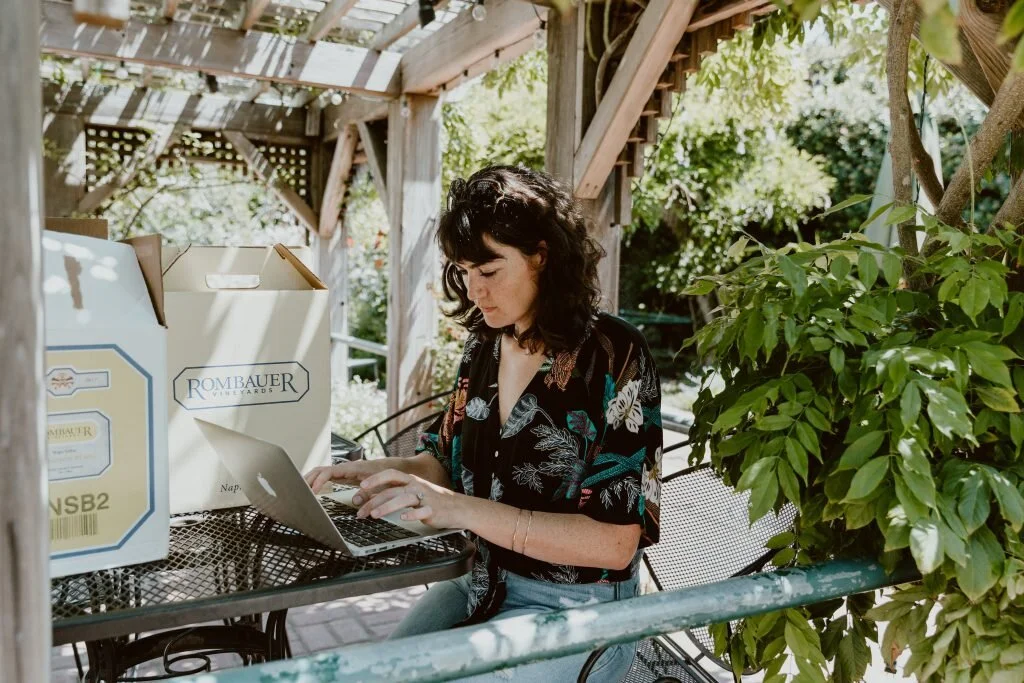 A woman with dark curly hair sitting at a metal table in an outdoor garden patio, working on a laptop, surrounded by greenery and wooden structures.