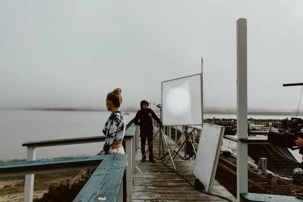 A woman stands on a wooden dock near a body of water, with a man operating photography equipment nearby and a large light reflector set up. The scene appears to be for a photoshoot.