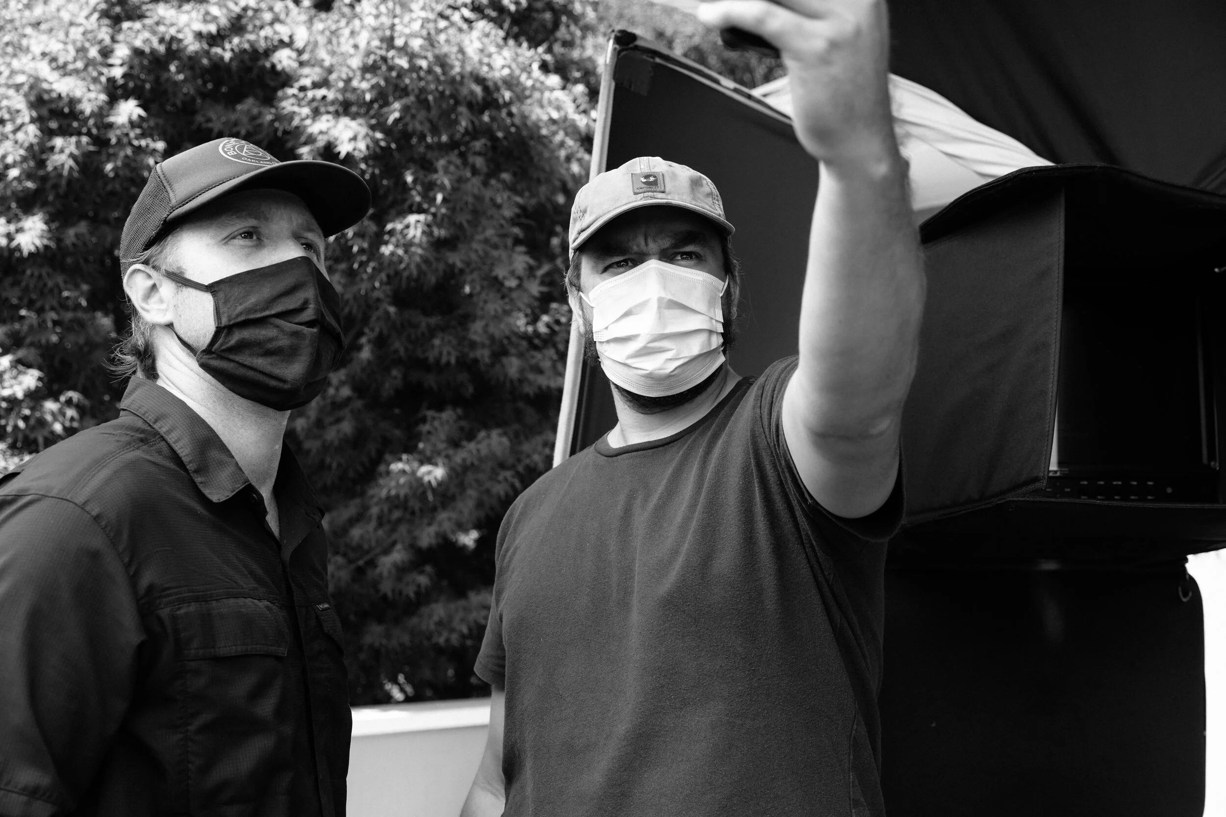 Two men wearing face masks and hats, one in a baseball cap and the other in a bucket hat, are standing outdoors in front of a leafy background. One man is looking at a screen or device, possibly a tablet, which he is holding and showing to the other 