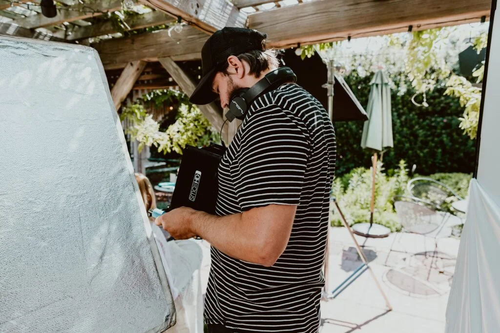 A man with a black cap, striped black-and-white shirt, and headphones around his neck, working with a laptop outdoors under a wooden shelter.