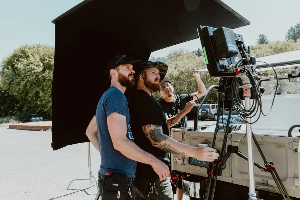Three men working on a film set outdoors, with one operating a camera and two others assisting, under a large black umbrella.