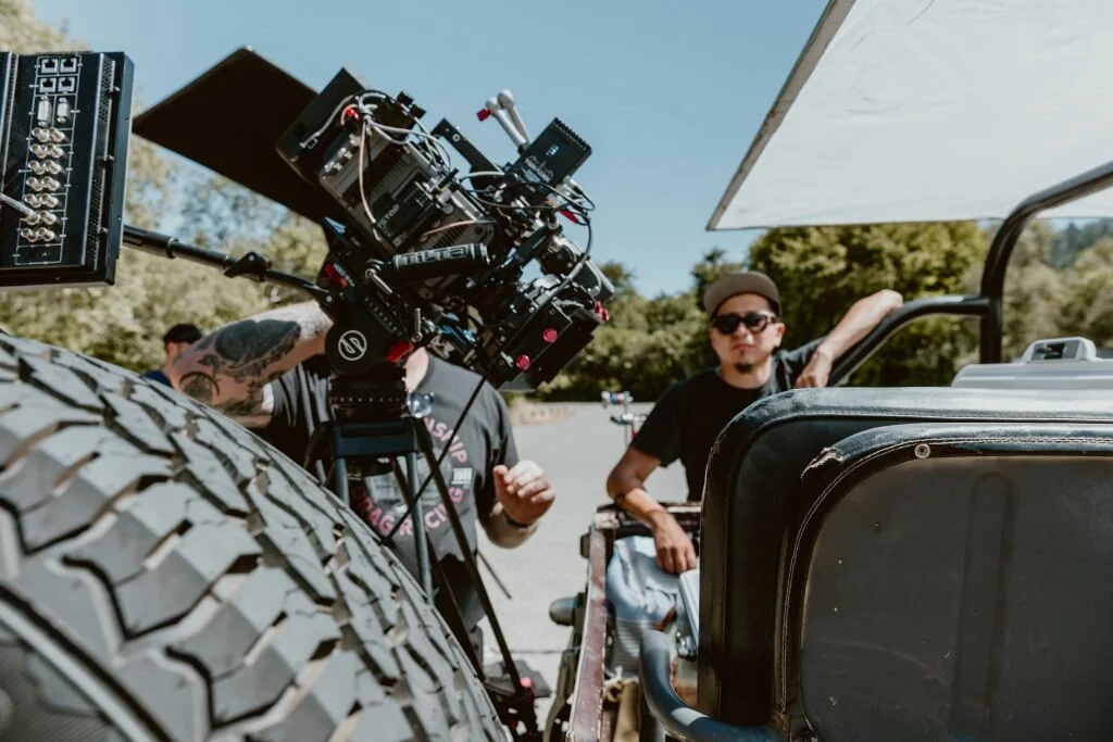 Filmmaking crew on set with camera equipment mounted on a vehicle, one person operating the camera and another standing nearby under a large umbrella.