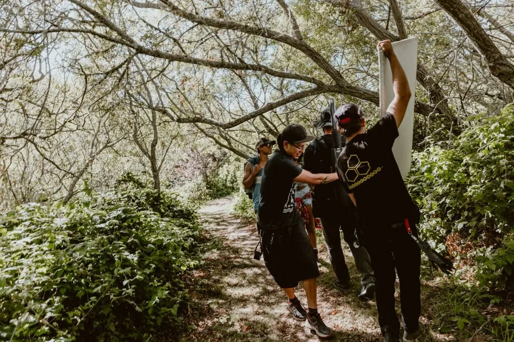 Group of people on a forest trail holding a large rectangular board or panel, setting up or adjusting it amid trees and foliage.