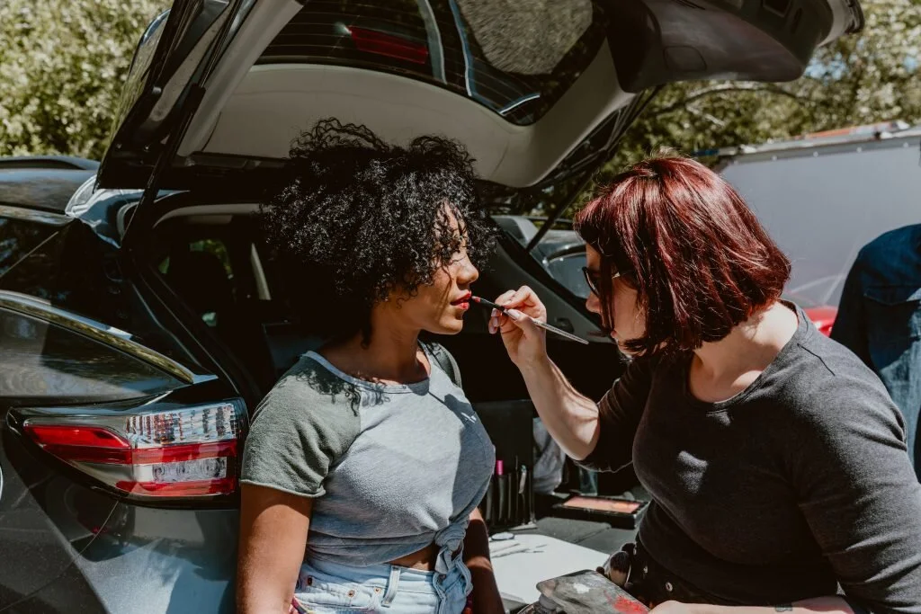 A woman with curly black hair is getting her face painted by another woman with red hair and glasses, outdoors near a black car with its hatch open.