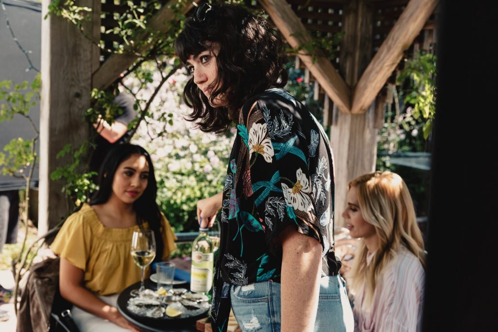 A woman with curly dark hair wearing a floral patterned shirt is serving food or drinks at an outdoor gathering with two women seated at a table with wine glasses, food, and lemon slices. The setting appears to be a garden or patio.