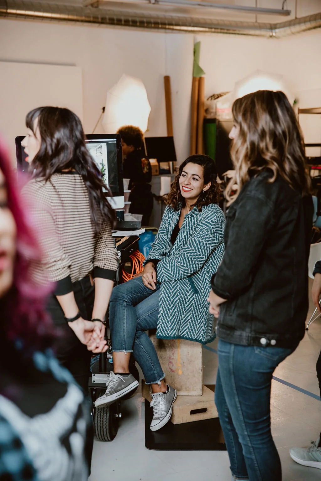Women conversing in a photo studio, one woman seated on a wooden box, smiling, surrounded by others in a creative workspace with studio lights and equipment in the background.