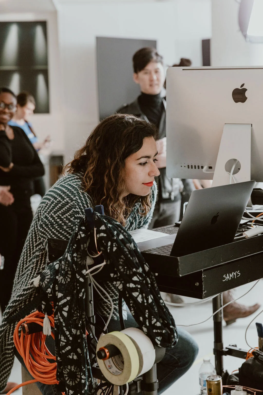 A woman with curly brown hair and red lipstick working on a computer at a desk, with a large monitor, surrounded by various electronic equipment and cables. Several people are in the background in an indoor setting.