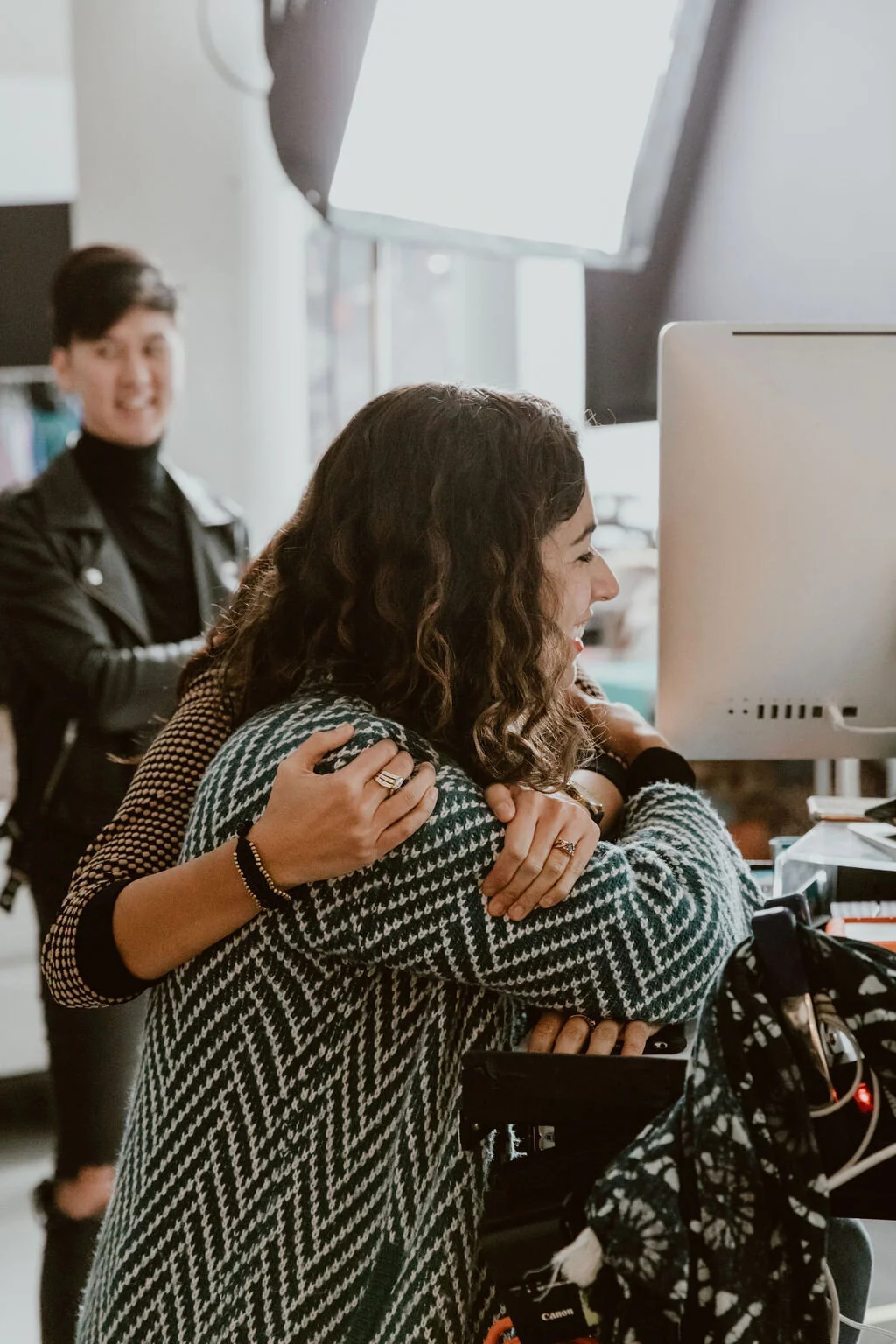 Two women smiling and hugging in front of a computer monitor in an office setting, with a third woman in the background.