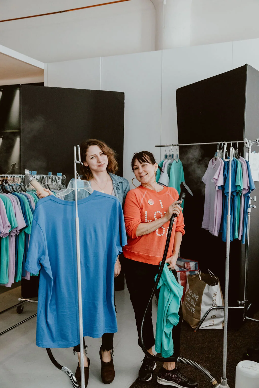 Two women standing in a clothing store with racks of colorful T-shirts.