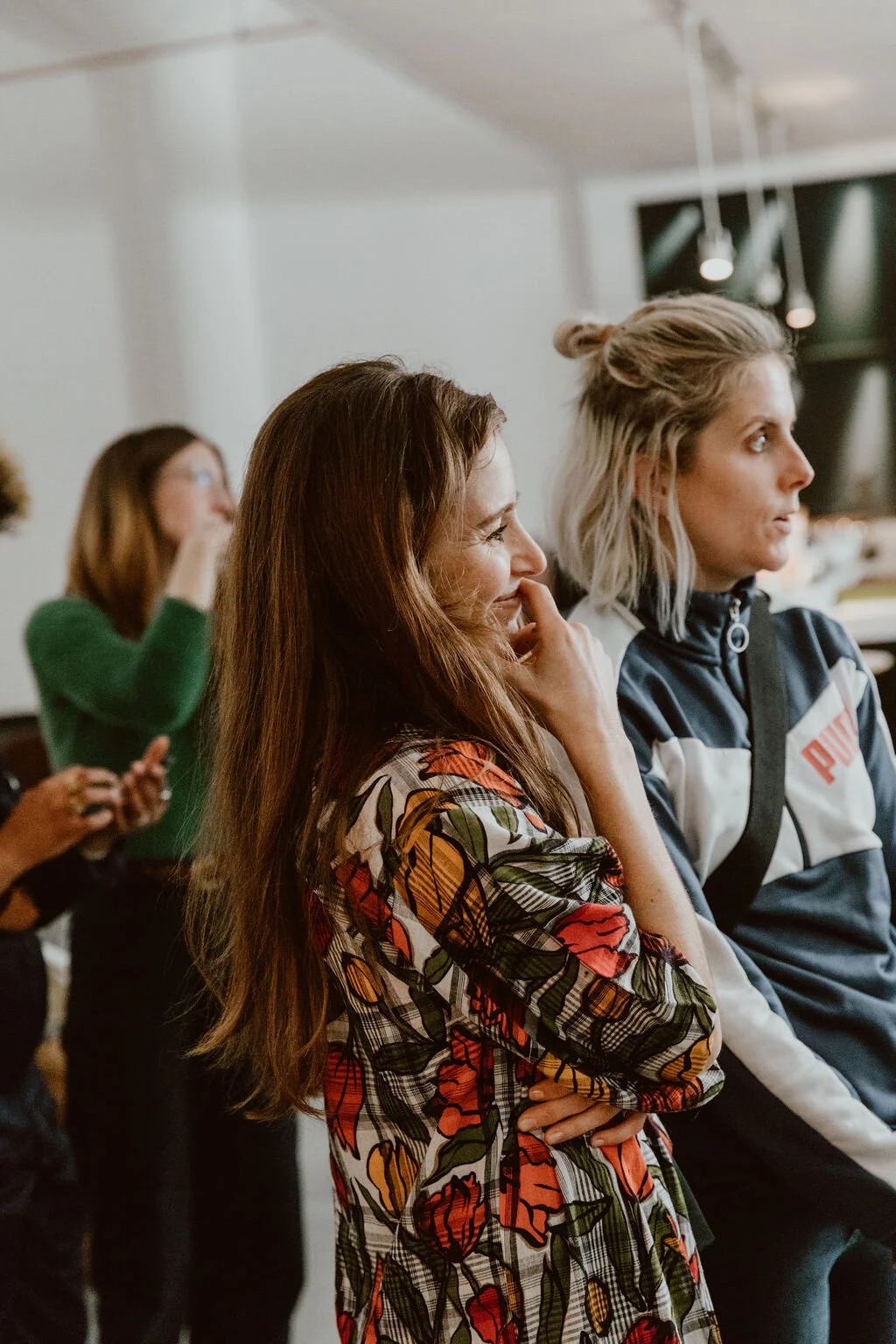 Two women standing in a room, listening attentively, with others in the background; one woman has long, red hair and is wearing a floral-patterned jacket, while the other has short, blonde hair and is wearing a black and white Puma jacket.
