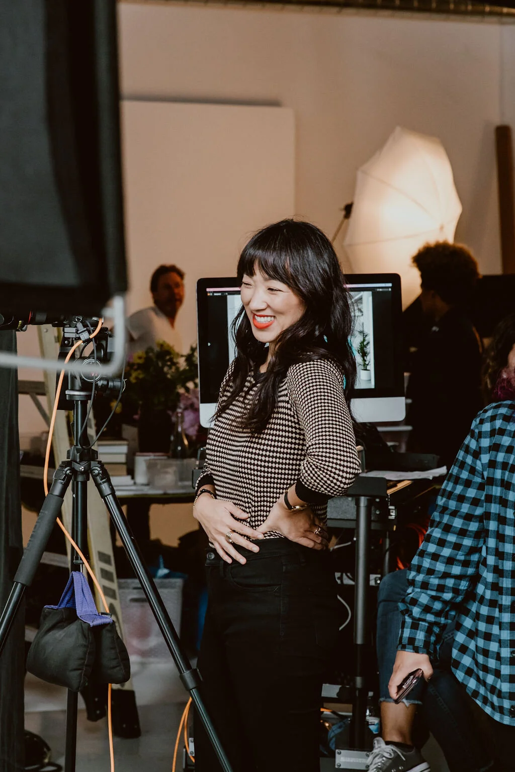 A woman with black hair and red lipstick smiling on a film set, surrounded by equipment and crew members.
