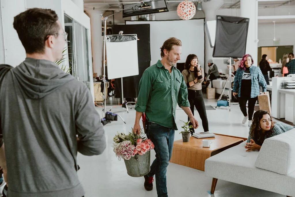 A man carrying a potted plant and a bucket of pink flowers walking in a studio with people and photography equipment.