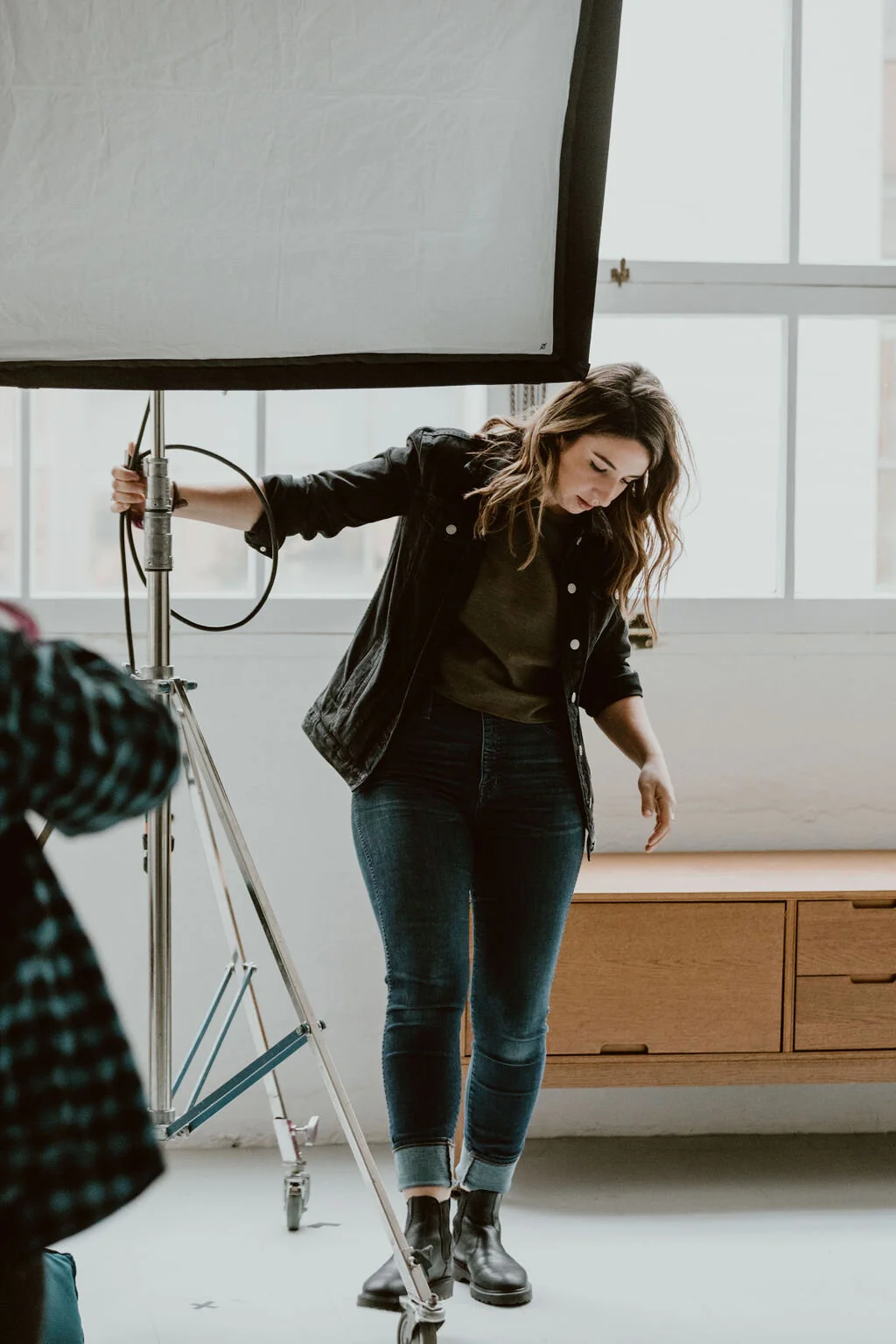 A woman with wavy brown hair wearing a black jacket, dark green shirt, blue jeans, and black boots standing in front of a large window during a photo shoot indoors, adjusting her position.