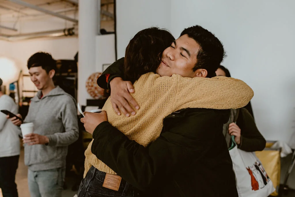 Two people embrace in a warm hug at an indoor gathering, with other people in the background.