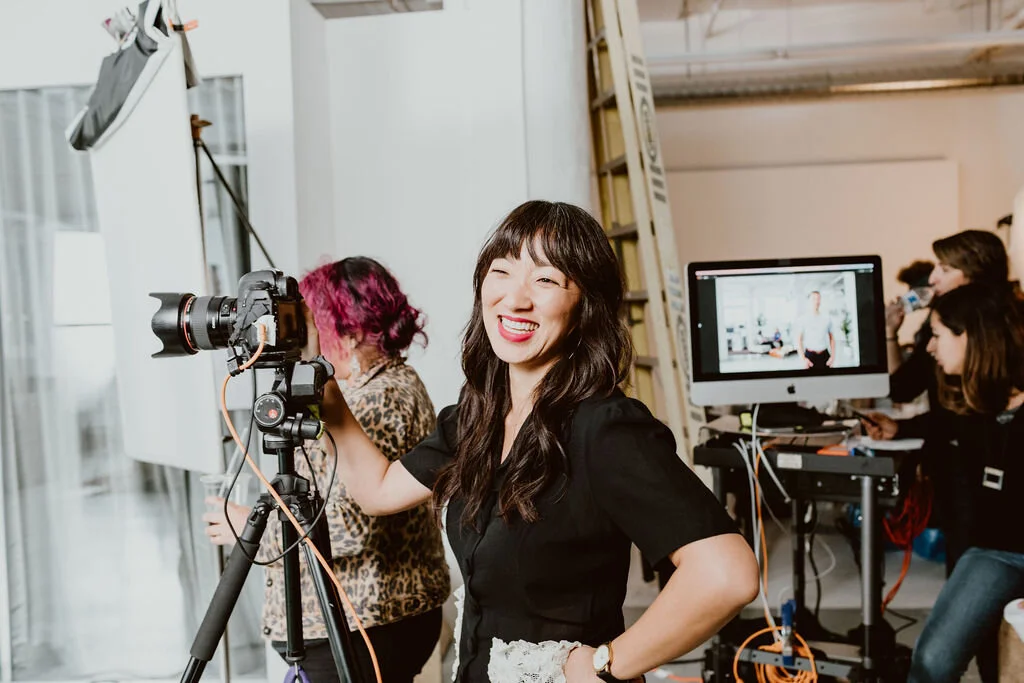 A group of people working in a photography studio with a woman smiling at the camera. The woman is operating a professional camera on a tripod. In the background, there is a large computer monitor and other individuals engaged in various tasks.