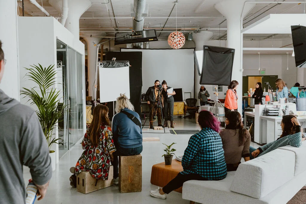 People attending a photo or video shoot in a large studio space, with equipment, lighting, and crew members preparing in the background.