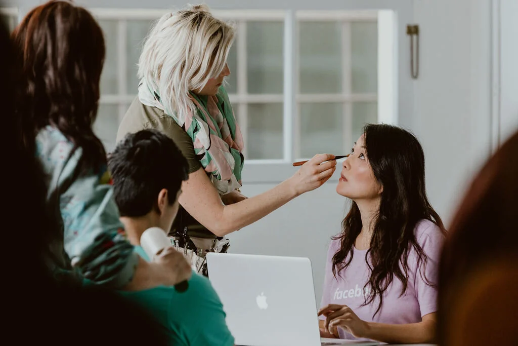 A woman is applying makeup to another woman seated at a table with a laptop in front of her, while several other people observe in the background.