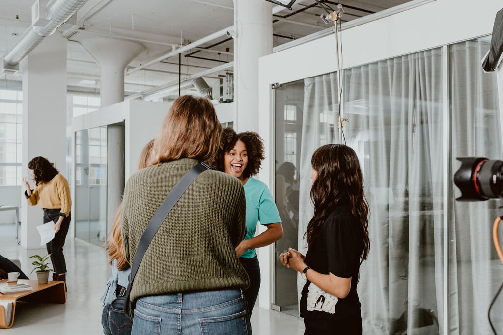 Group of four women having a conversation in a modern office space, with one woman in the background talking on her phone.