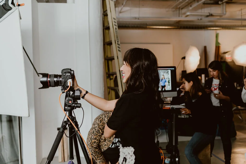 A woman in black is adjusting a camera on a tripod during a photoshoot in a studio with team members working at computers in the background.