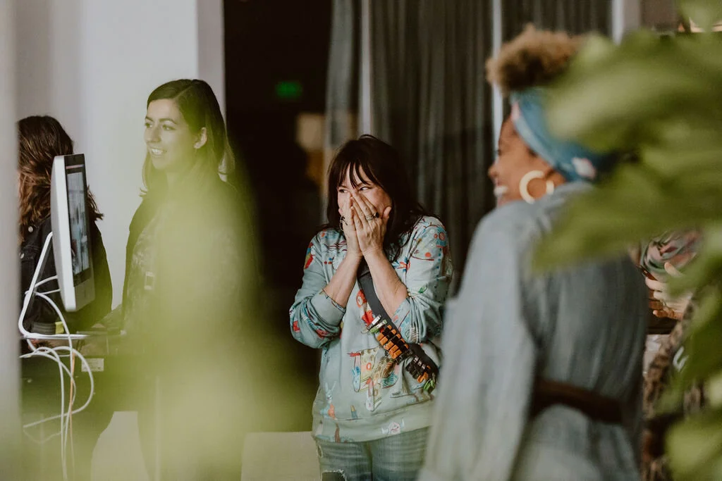 Women in an office sharing a joyful moment, some are smiling and laughing, with one woman covering her mouth in surprise.