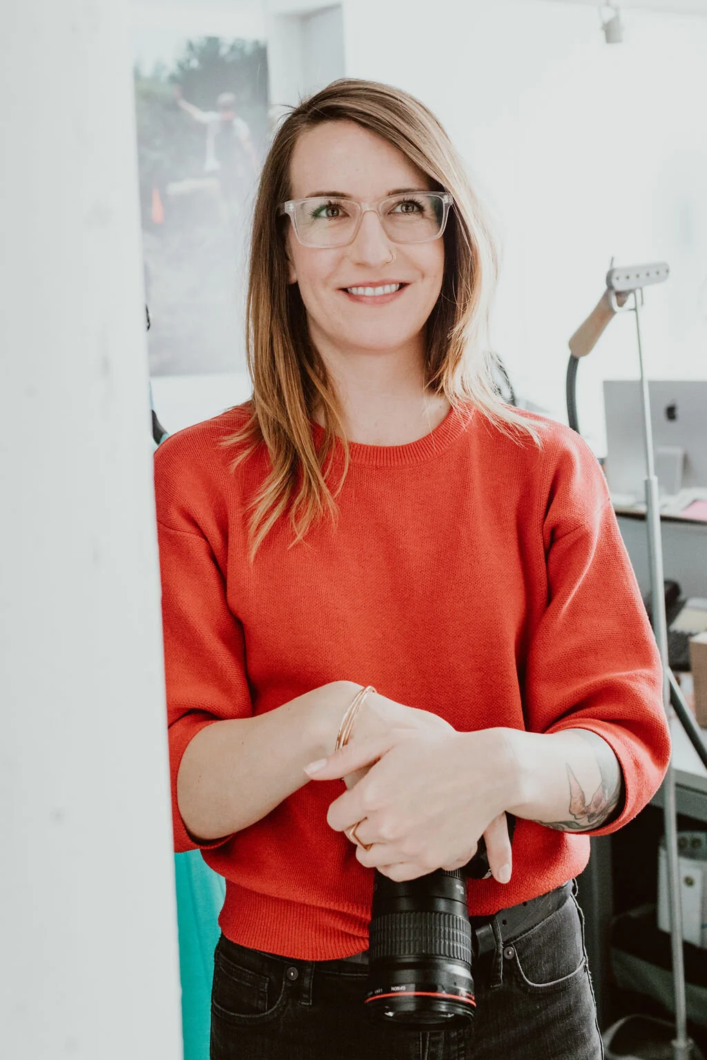 A woman with shoulder-length light brown hair wearing glasses and a red sweatshirt, holding a camera, and smiling.