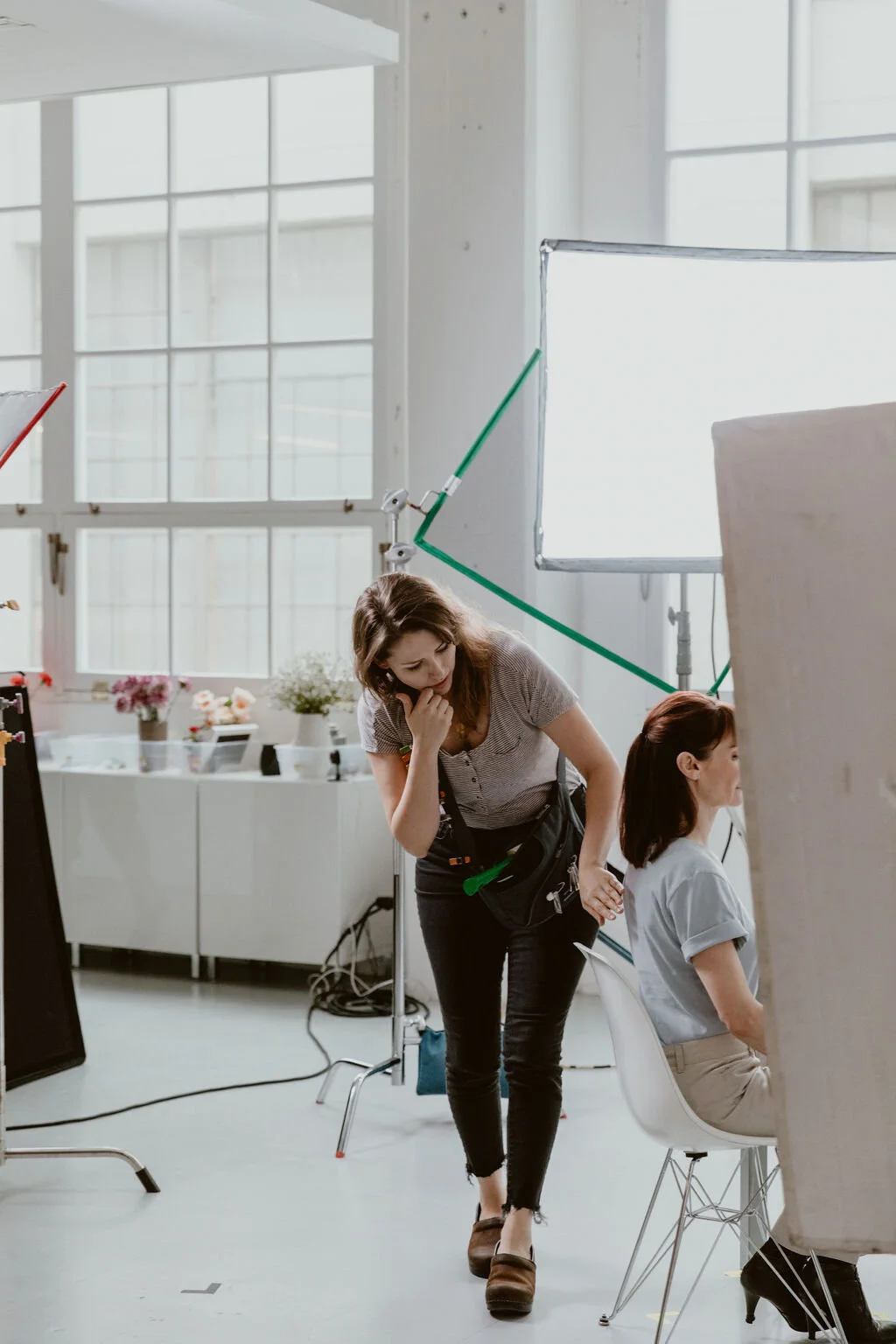 A woman is standing next to a seated woman in a bright, airy studio with large windows and studio lighting equipment around them.