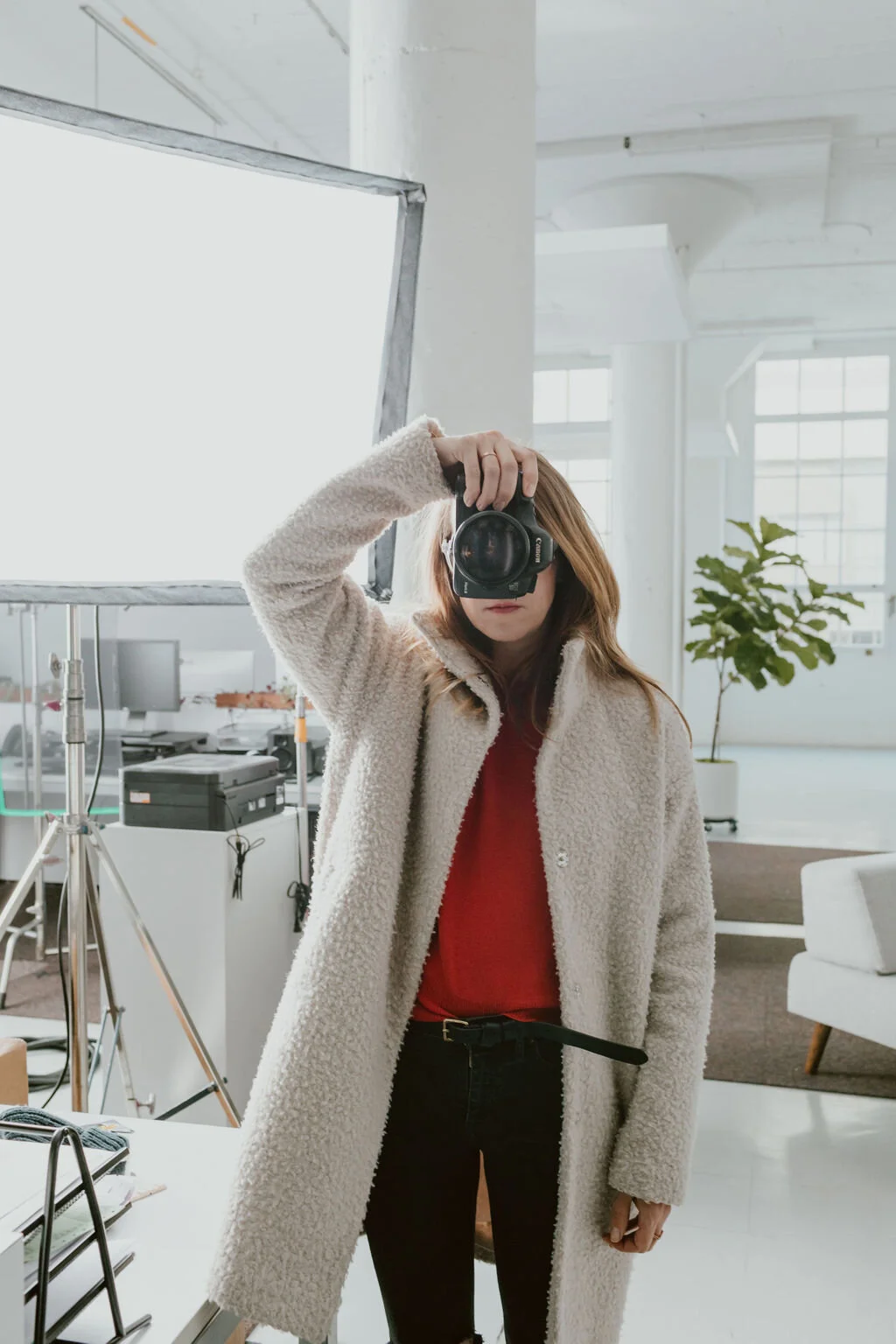 A woman taking a selfie with a camera in a modern office or studio space. She is wearing a beige coat and red shirt, with a potted plant and large windows in the background.