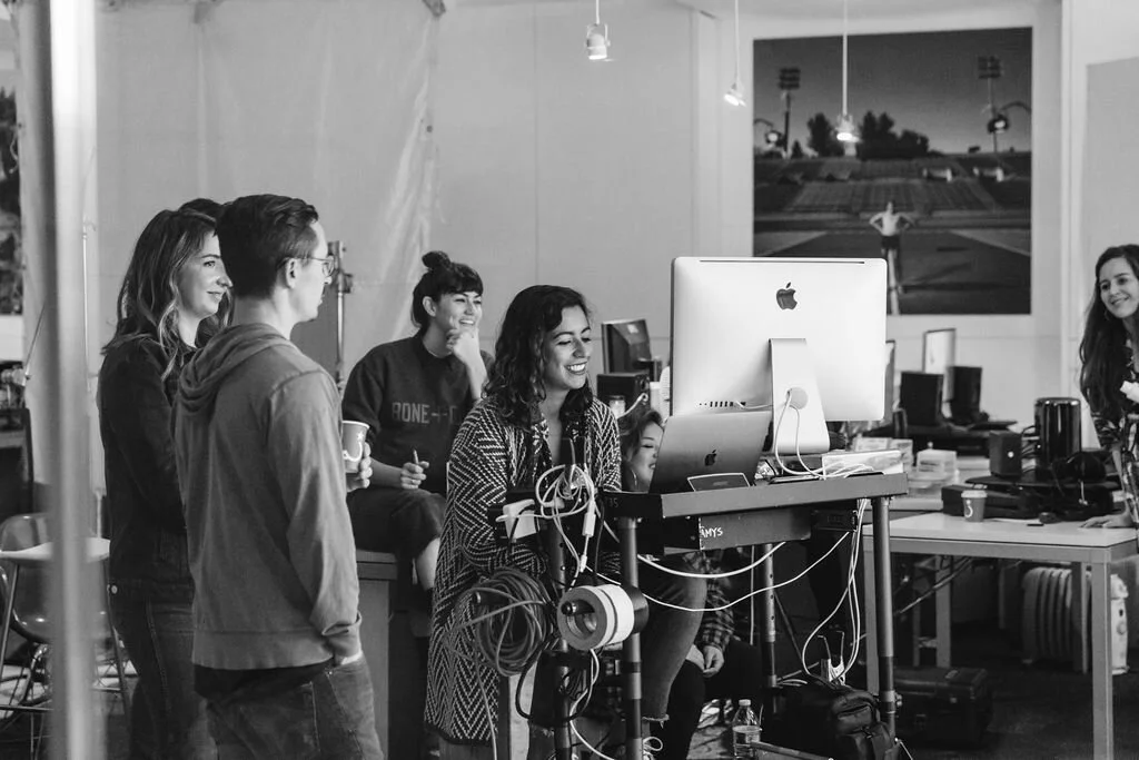 Group of people in a studio working with computer on a project, some smiling and chatting, with equipment and monitors around.