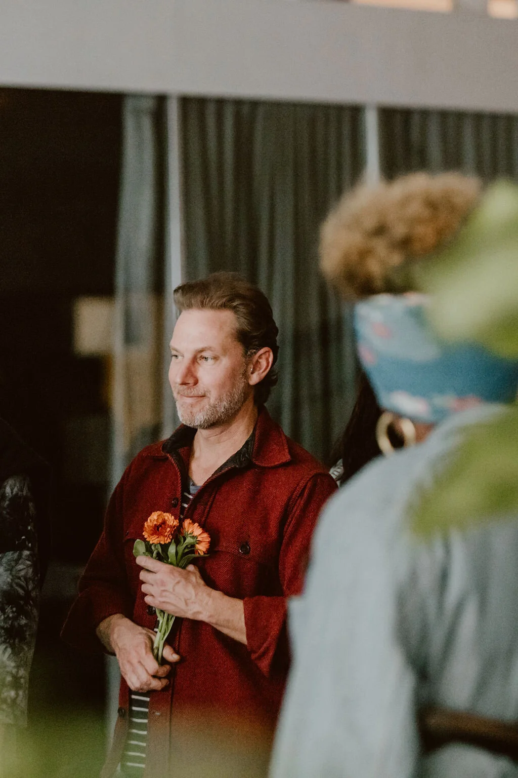 A man with a beard and mustache wearing a red jacket holding orange flowers, standing indoors with dark curtains in the background.