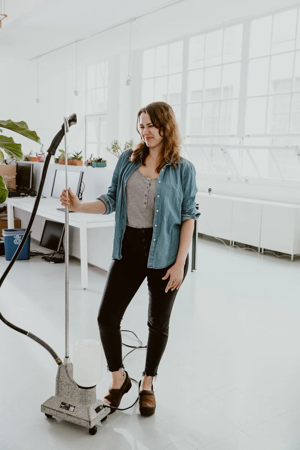 A woman in casual attire, standing in a bright room, looks uncomfortable or distressed while touching a fogger machine.