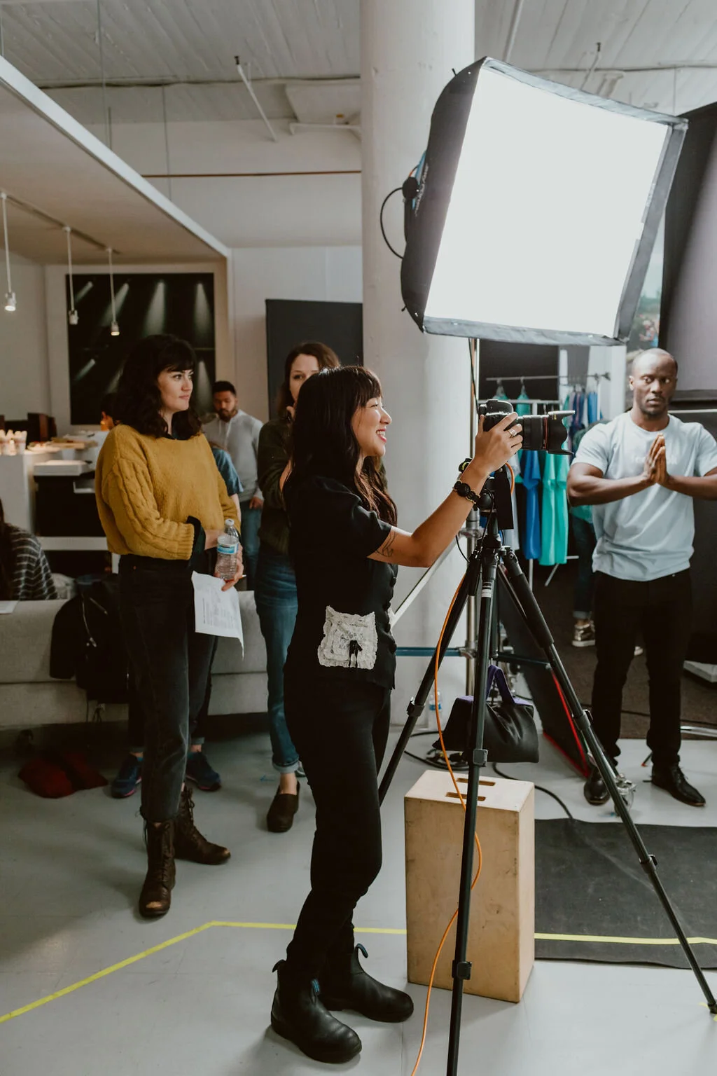 A group of people in a creative space, with a woman operating a camera on a tripod, standing near a large light diffuser. Others are watching, with some holding water bottles and papers, and a man in a white shirt with hands in prayer position.