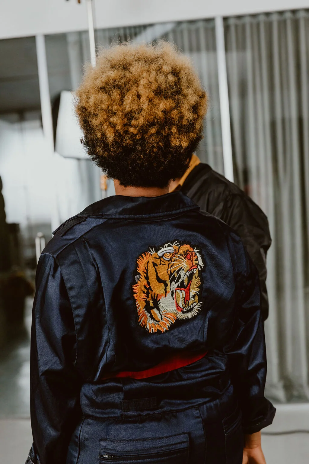 Back view of a person with curly blonde hair wearing a navy blue satin bomber jacket with an embroidered tiger head on the back, in an indoor setting.