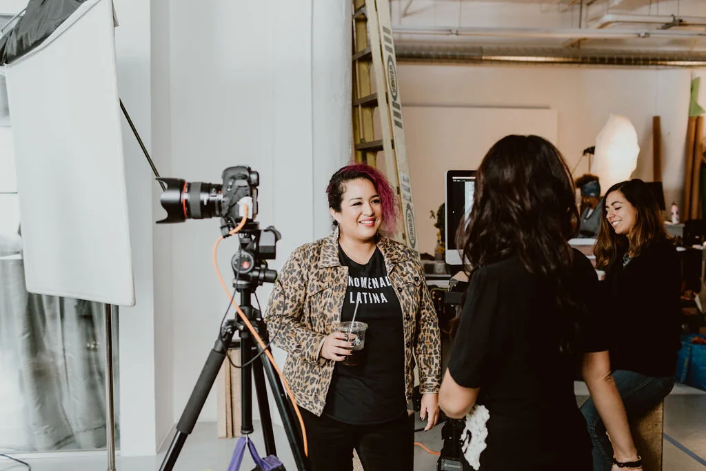 A woman with pink and black hair smiling and holding a drink in a photography studio, with photographers and photography equipment around her.