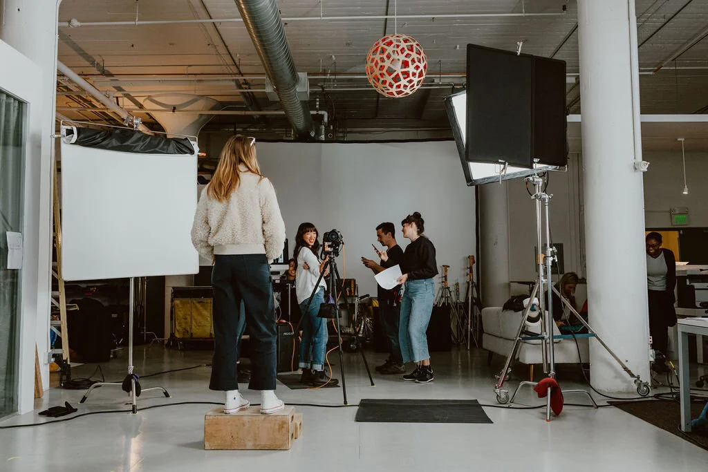A video production crew setting up equipment in a studio, including a camera on a tripod, lighting, and a backdrop, with several crew members preparing for filming.
